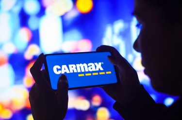 Person holding a smartphone displaying the CarMax logo on the screen against a blurred, colorful background.