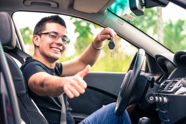 Smiling man sitting in the driver’s seat of a car, holding up a set of car keys and giving a thumbs-up gesture.