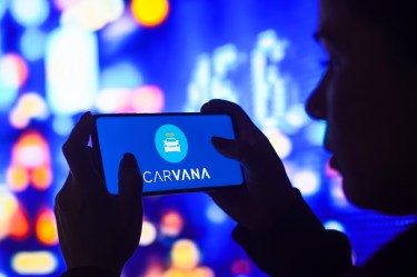 Person holding a smartphone displaying the Carvana logo on the screen against a blurred, colorful background.