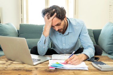 Man reviewing bills at a table with a laptop and calculator, holding his head in stress while going over paperwork.