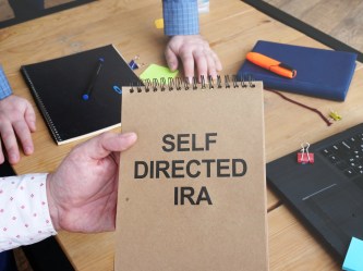 Notebook labeled “Self Directed IRA” on a desk with financial materials and a laptop.