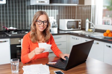 Woman sitting at a laptop at a kitchen table, holding receipts