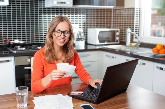 Woman sitting at a laptop at a kitchen table, holding receipts